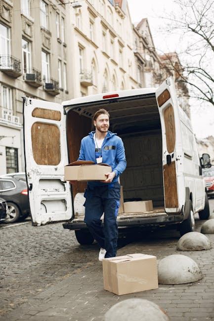 A man with dark skin, wearing a black quilted vest over a blue long-sleeve shirt, black trousers, white sneakers, and a white cap, stands beside an open white van with a wooden panel on the side durin